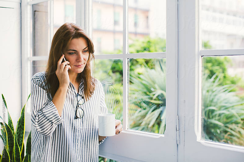 Woman holding a coffee mug talking on phone by window at home.