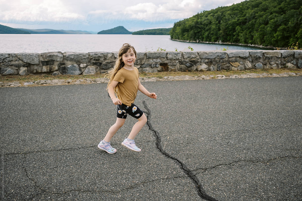 smiling girl running on road in front of beautiful lake and rock wall