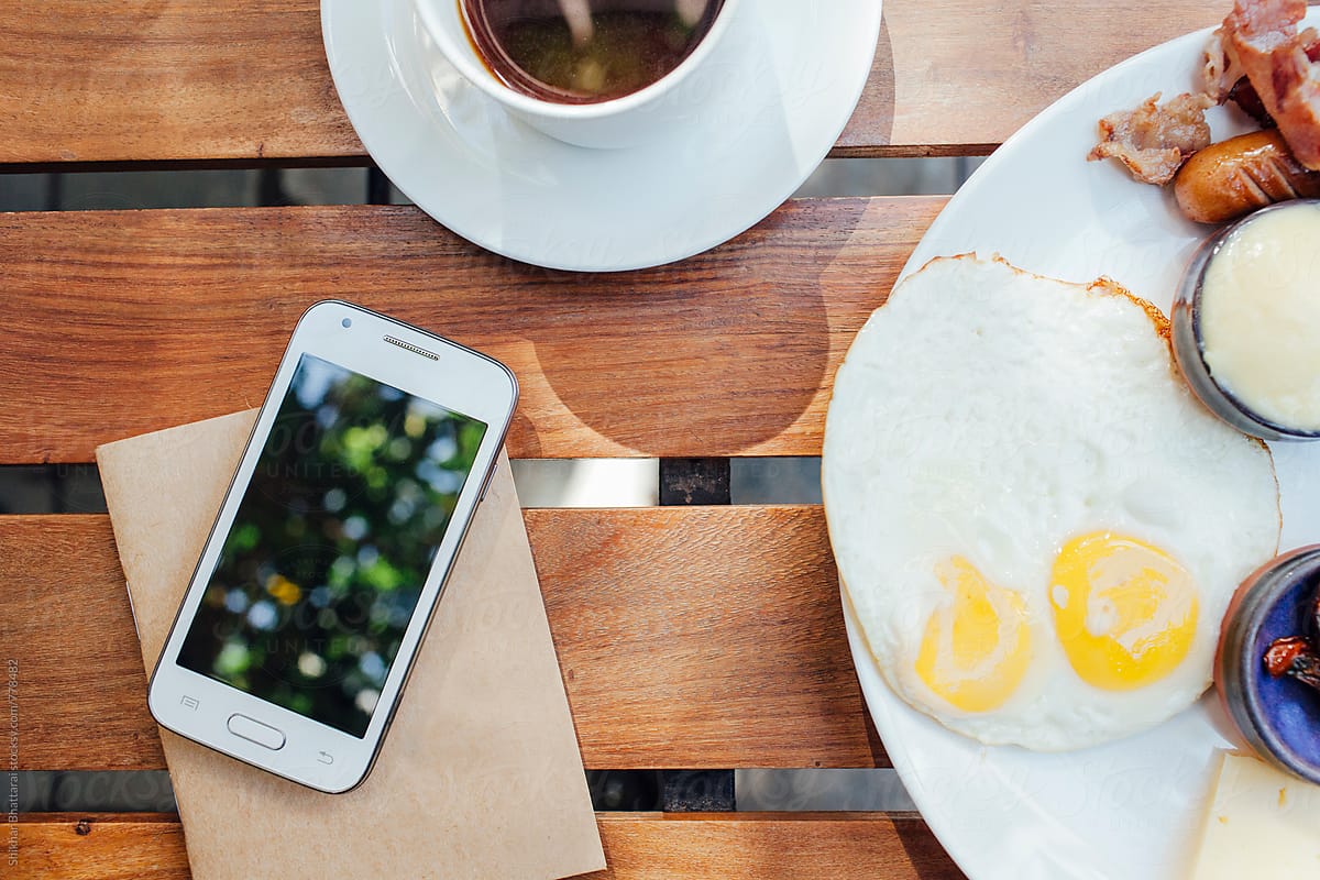 Blank cell phone on top of a notebook with breakfast on the side.