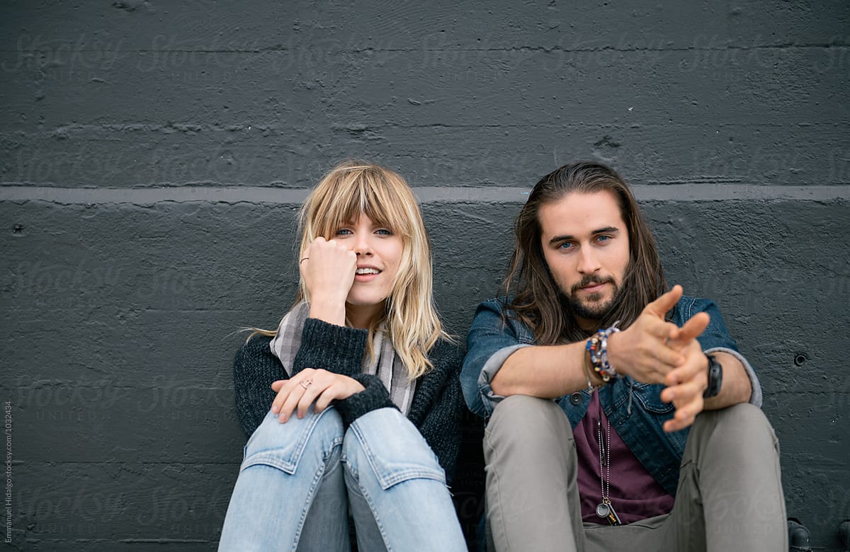 Young attractive couple sitting against wall and looking at camera