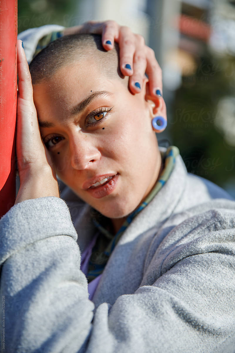 Depressed woman with shaved head leaning on metal bar in city