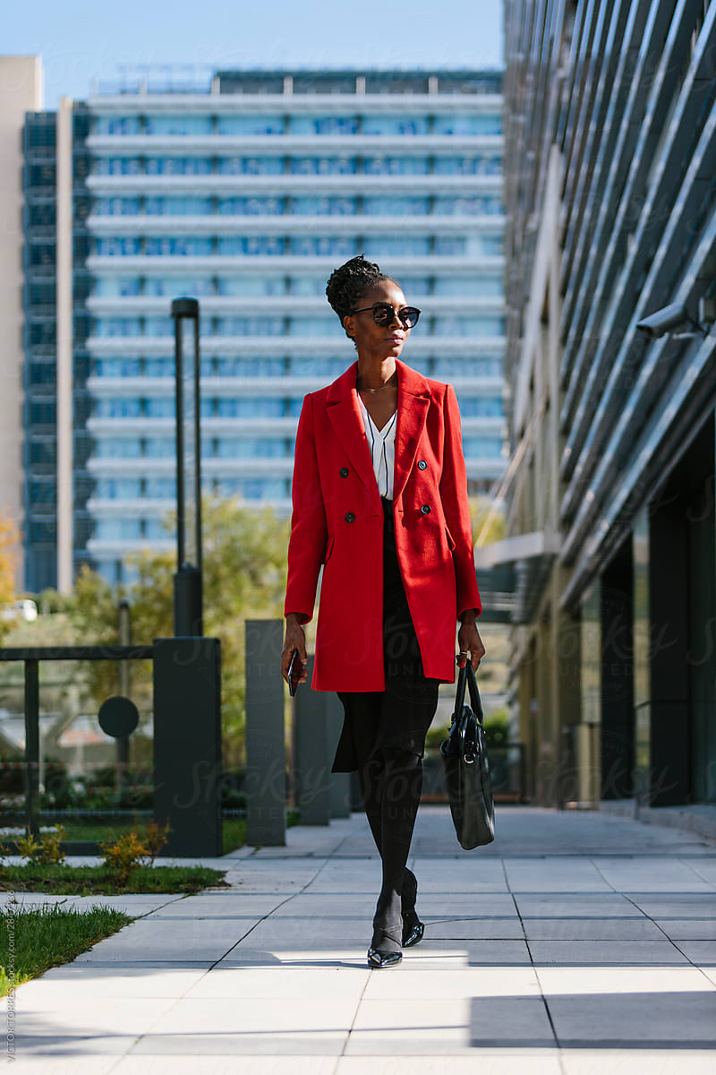 Fashionable black woman in red coat walking along glass buildings in downtown
