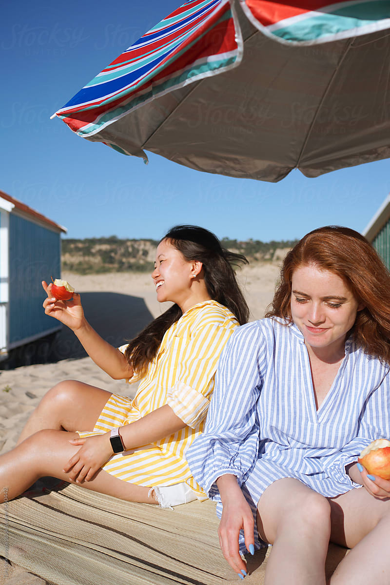 Two girls sitting on the beach laughing and eating apples