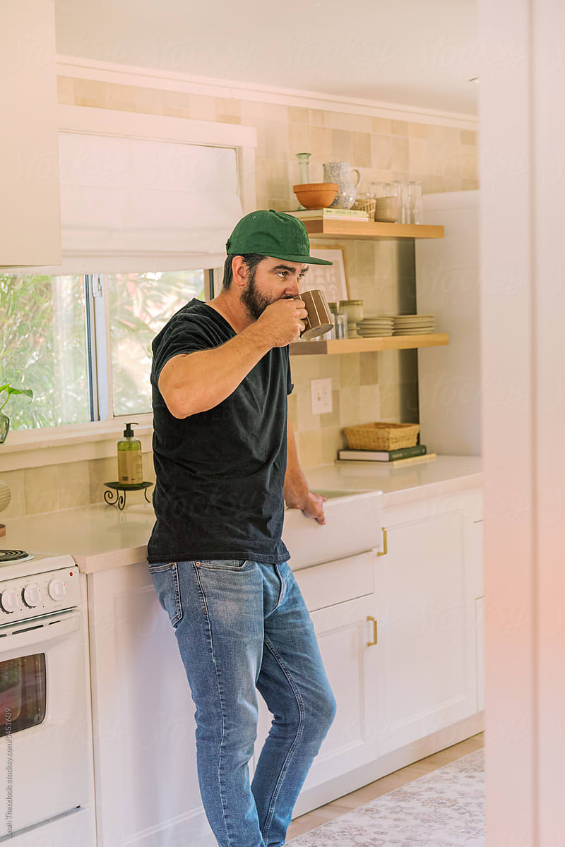 Millennial bearded man in kitchen, sipping a cup of coffee