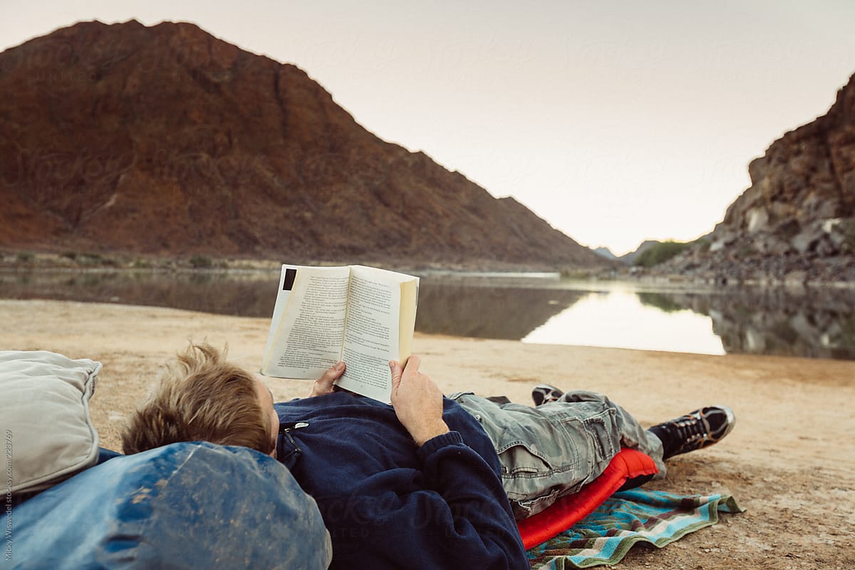 Man lying at his overnight camp next to the river reading a book