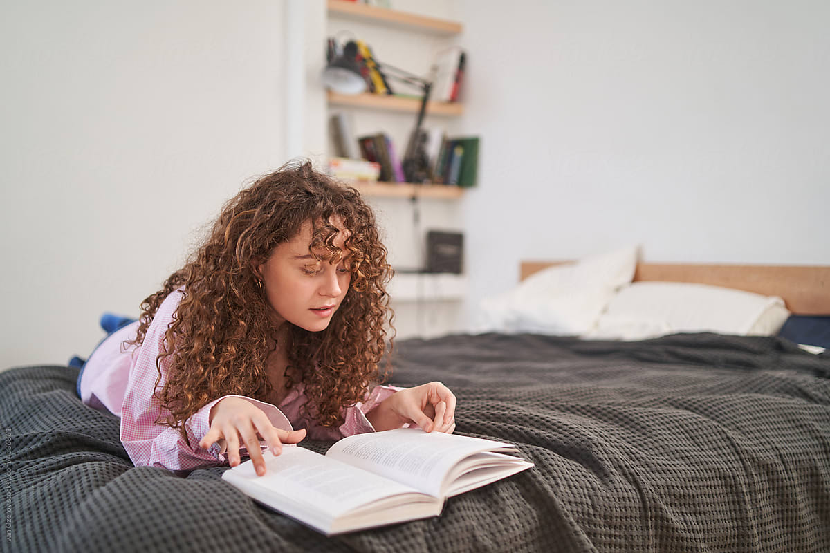 curly young woman reading book lying on bed