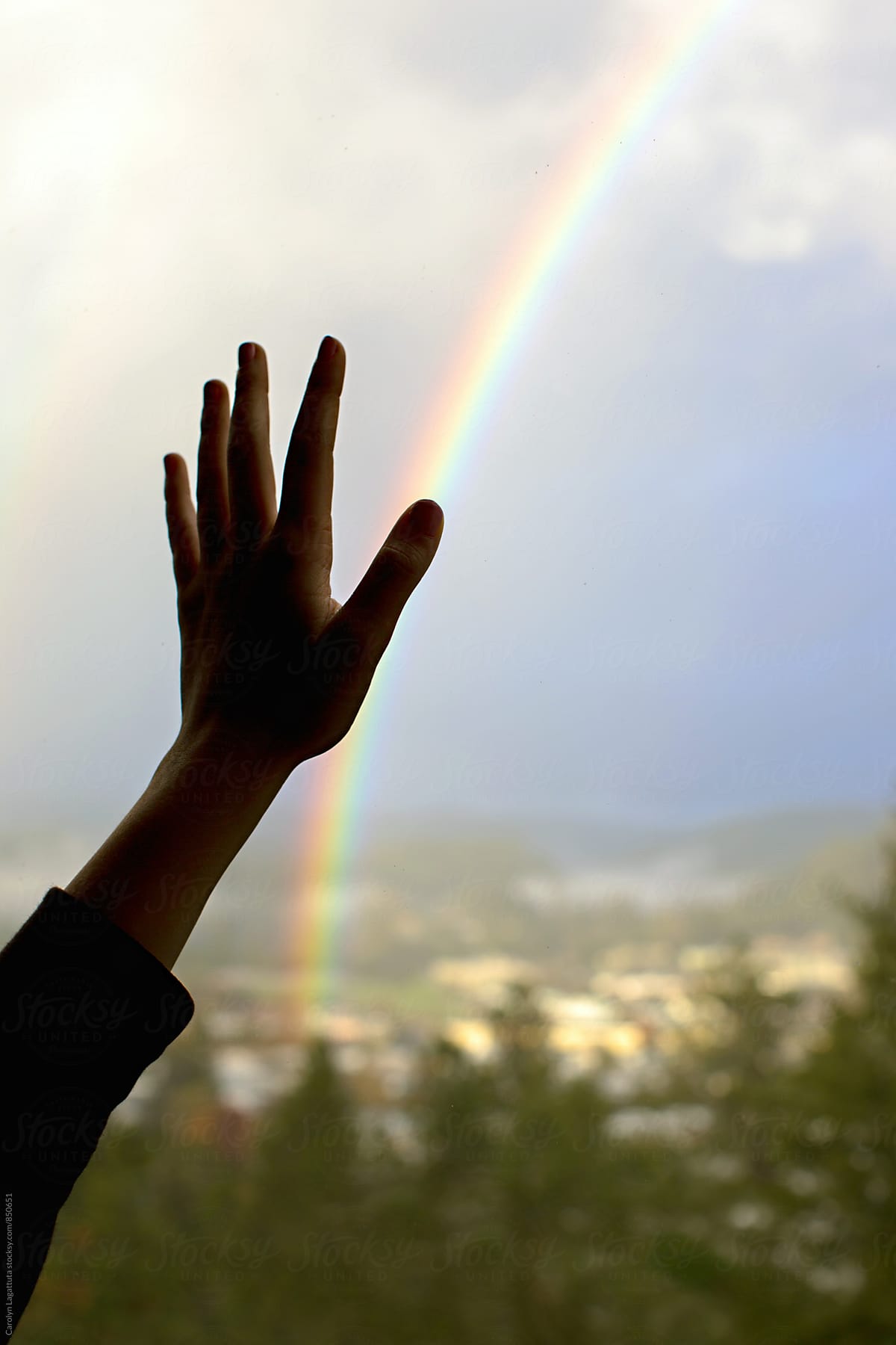 Hand pressed up against a window with a view of a valley and a bright rainbow