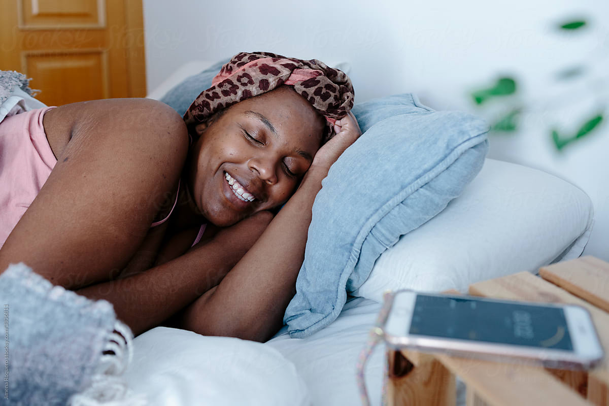 Woman in bed smiling lazily while alarm clock rings on cell phone