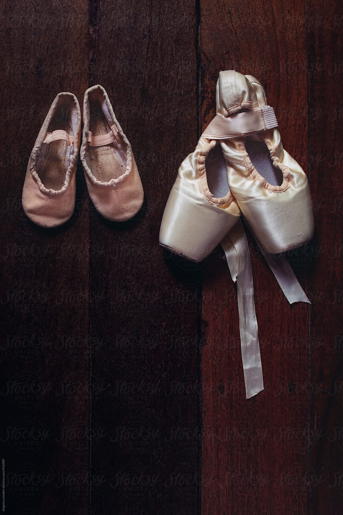A small child's old ballet shoes beside a pair of pointe shoes on a dark wood background