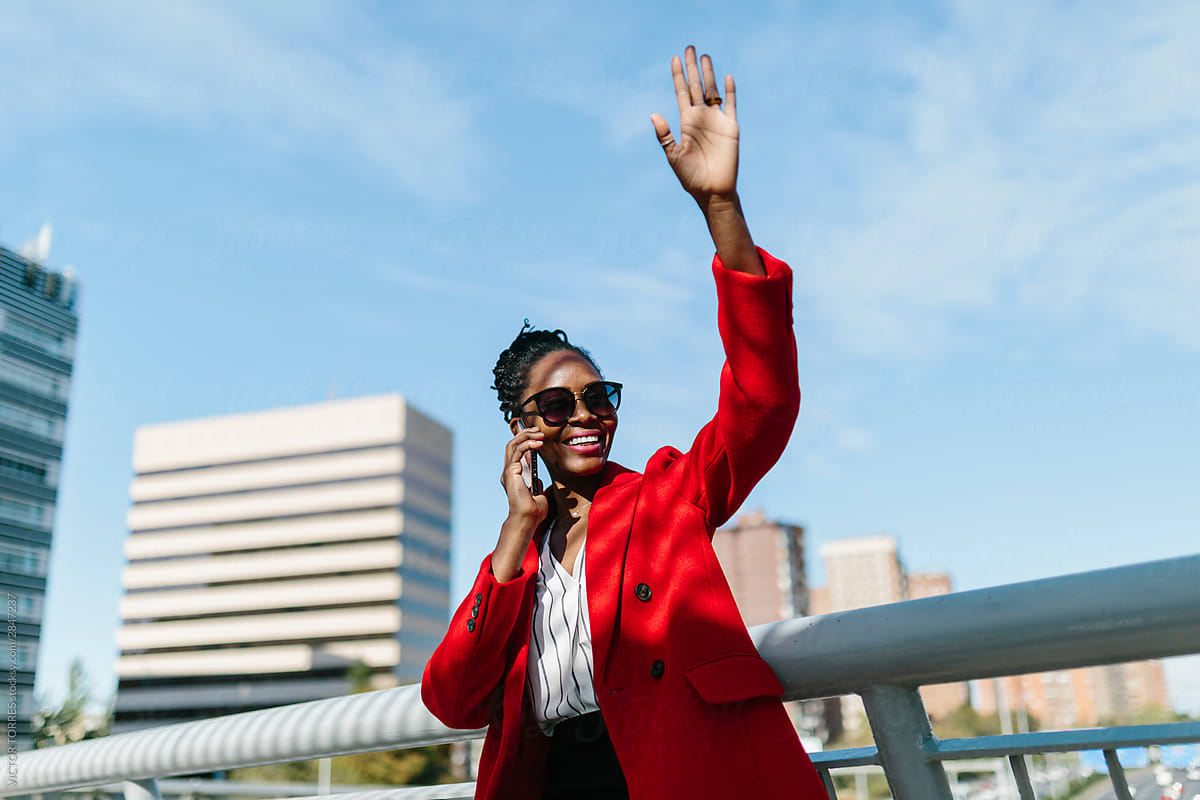 Smiling black woman in red jacket talking on smartphone and raising hand