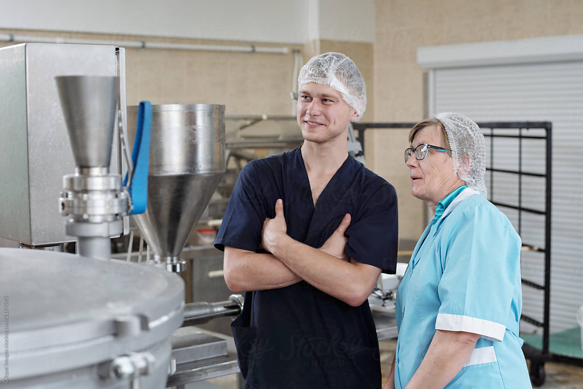 Young male manufacturing worker standing at food processing plant with elderly female colleague, looking at modern machinery and smiling
