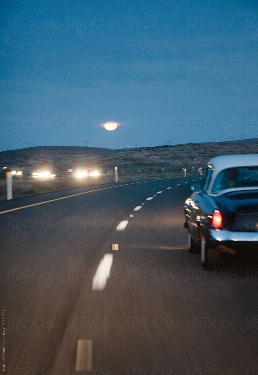 Vintage car driving on highway toward moon at twilight