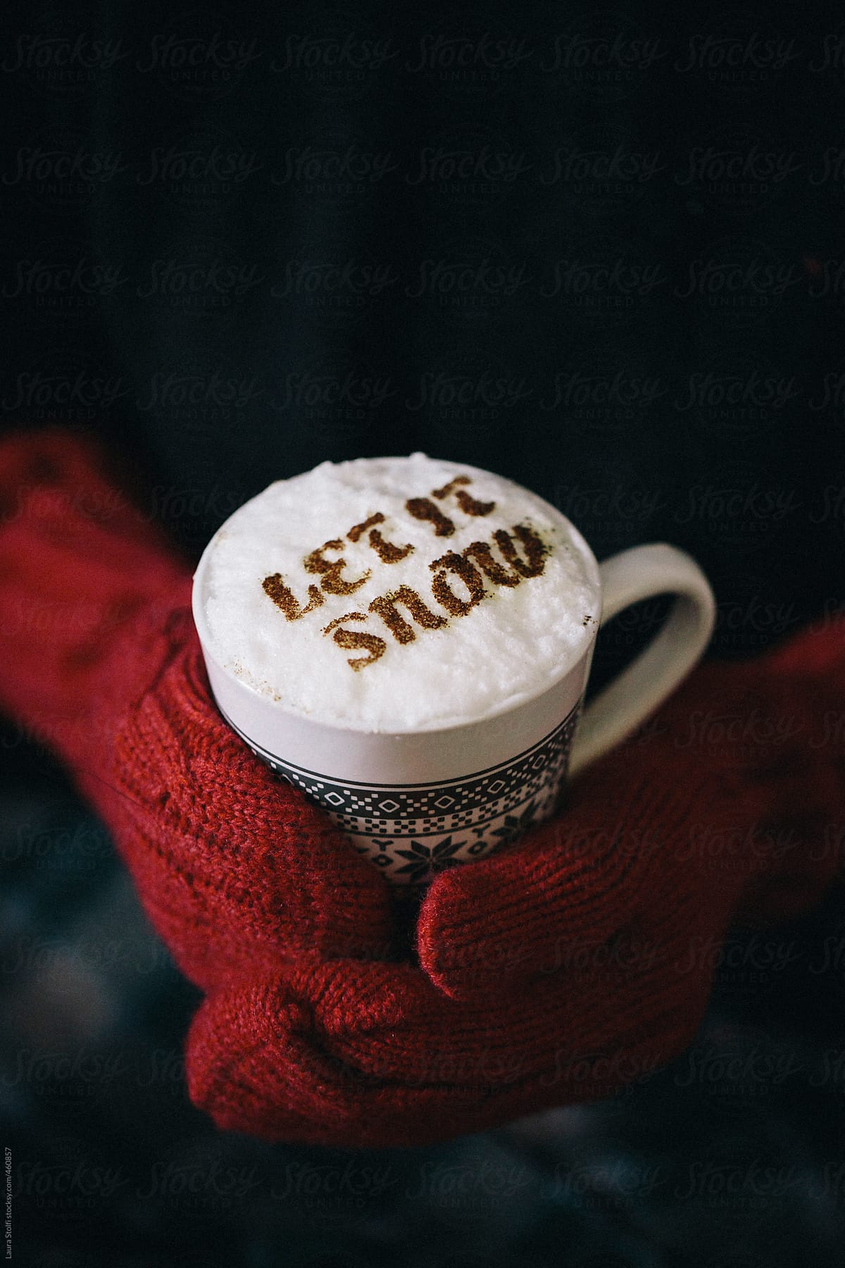 Hand with red mittens holds a cup of cappuccino with write Let it snow on top