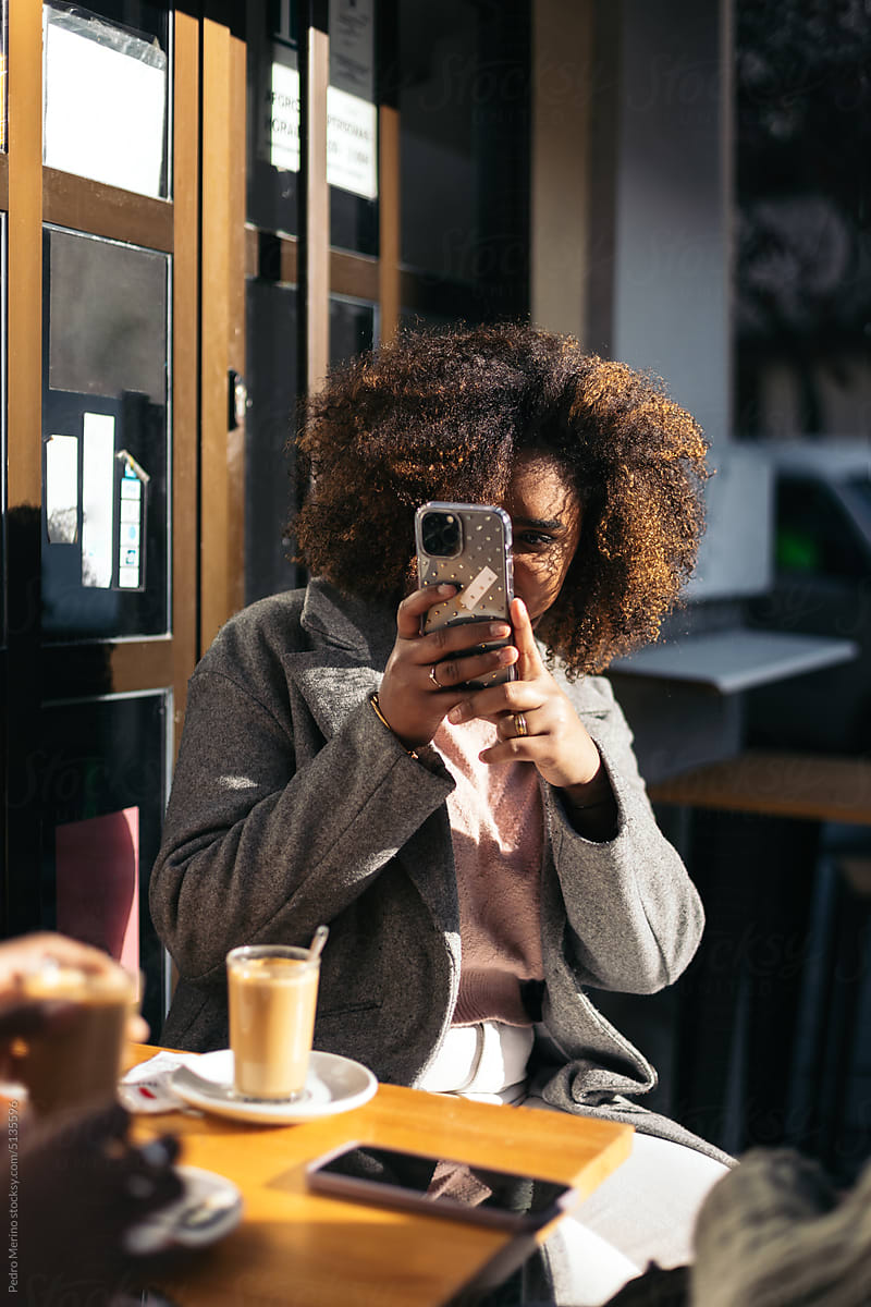 Woman with afro hair taking photos with smartphone drinking coffee