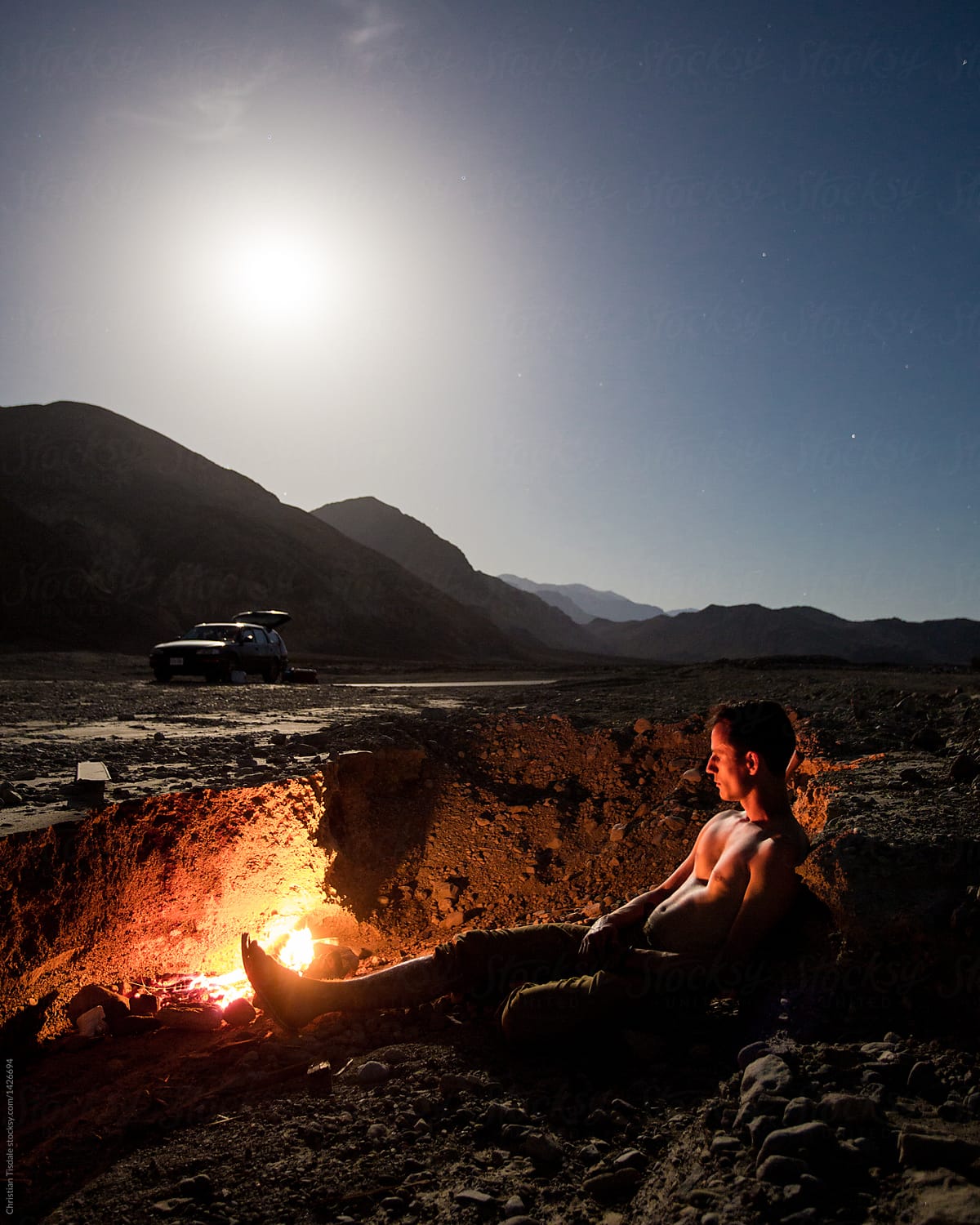 A young man sitting beside a fire under the moonlight in the desert