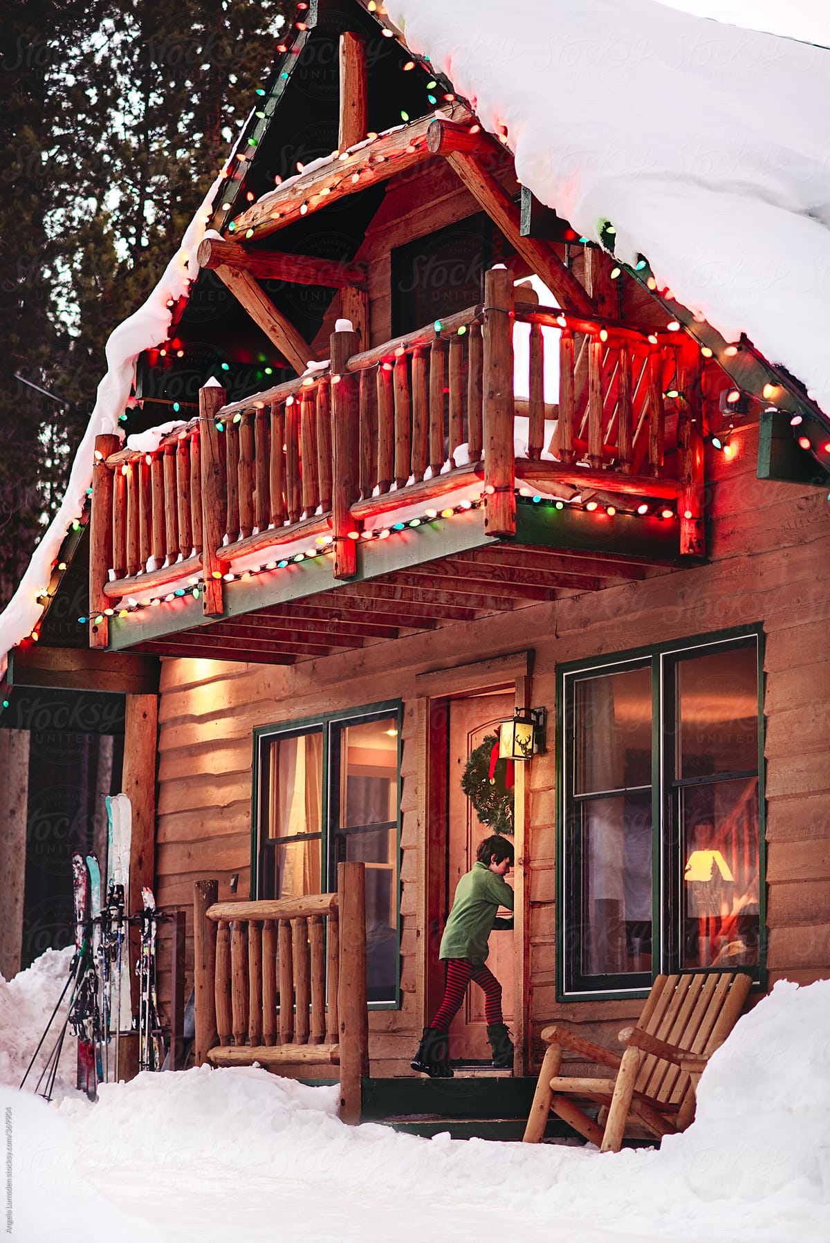 Boy goes in the front door of a cabin decorated with holiday lights at dusk in snow