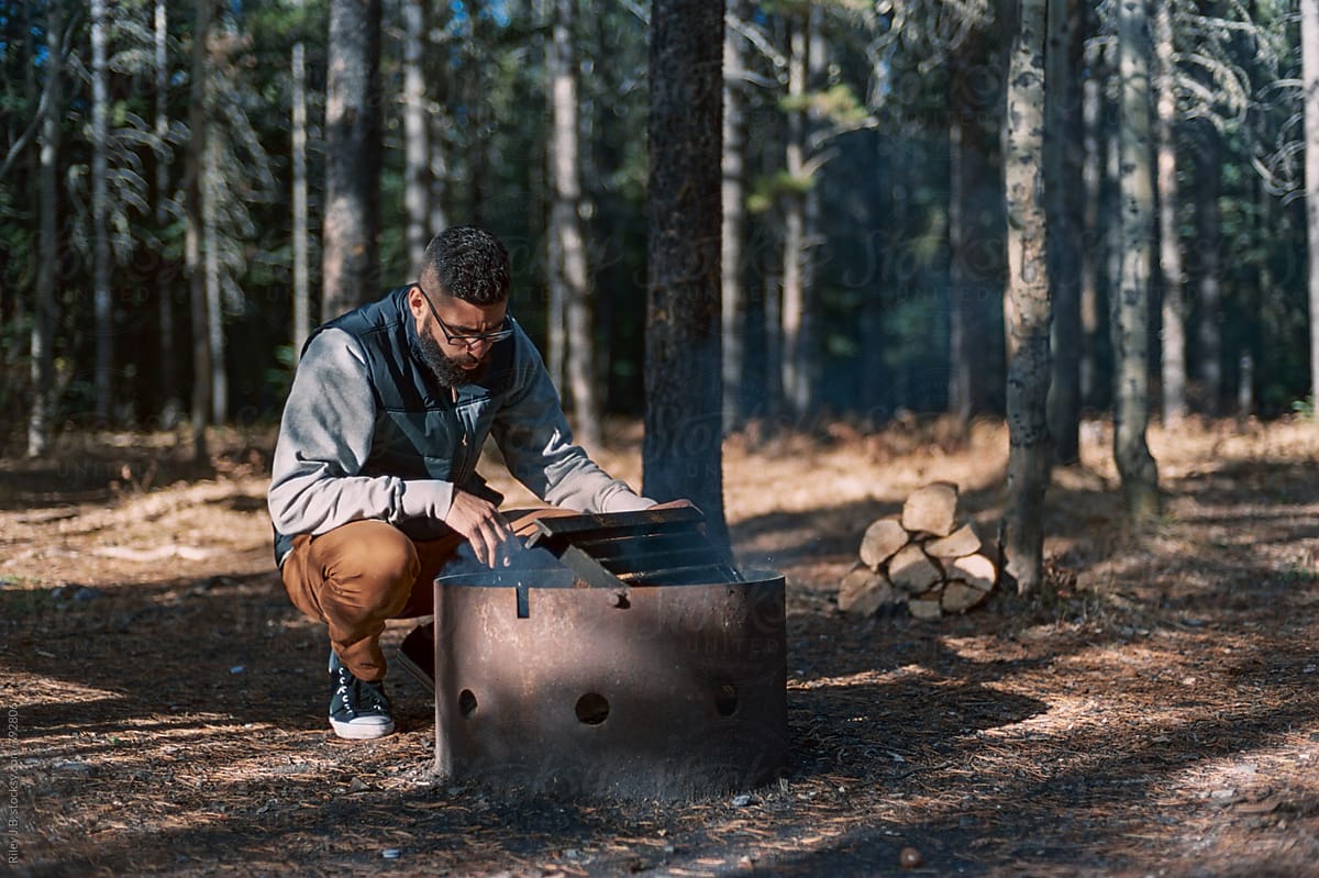 A young bearded man tends to a fire pit while camping.