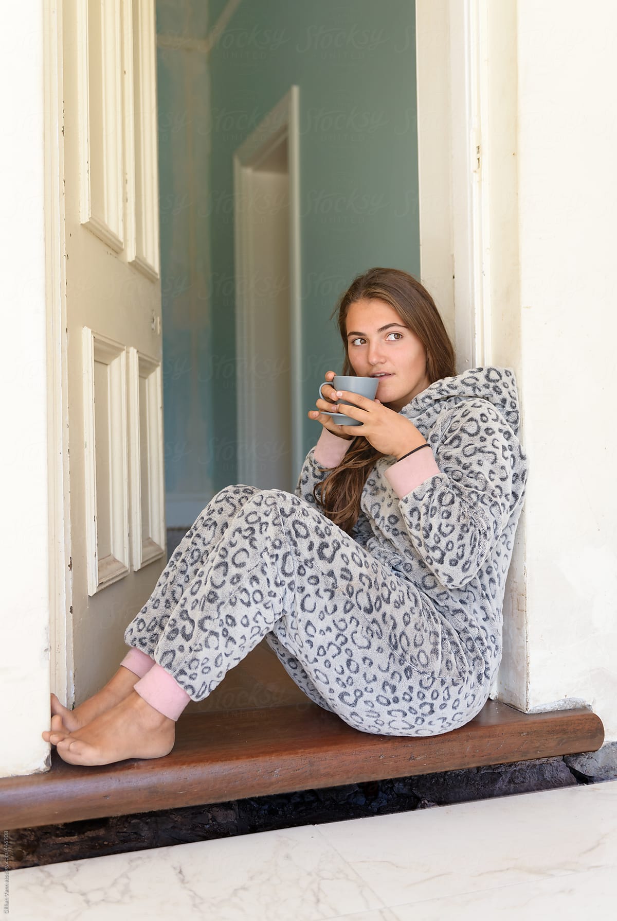 Young Woman Wearing A Onesie Pyjama Drinking Tea Or Coffee In The Doorway" by Contributor "Gillian Vann" - Stocksy
