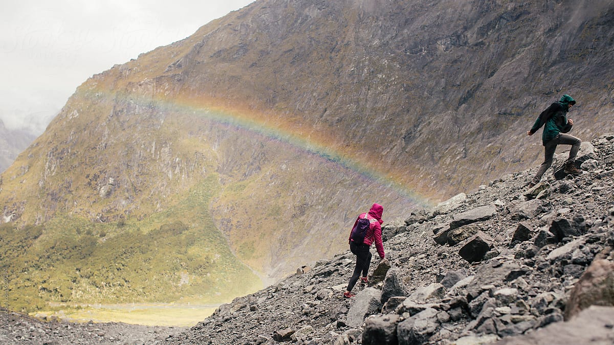A couple of hikers climbs a rocky mountain with a rainbow in the background