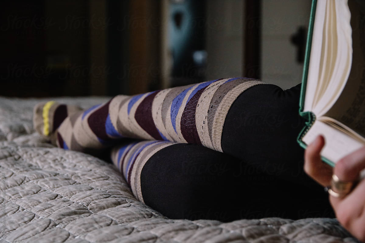 Woman holding a book laying on a bed wearing argyle pattern knee-high socks