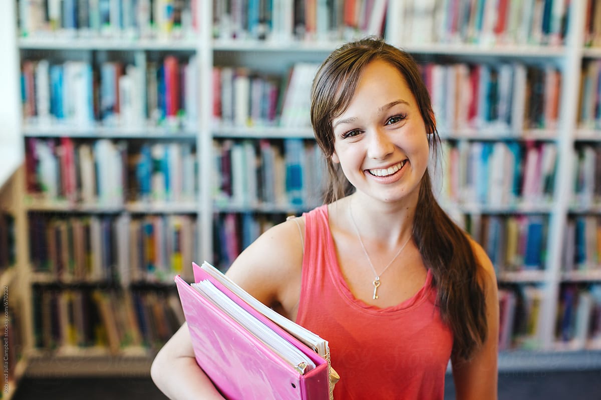 Portrait Of Happy High School Teenage Girl Holding Book Binder In Library  by Rob And Julia Campbell