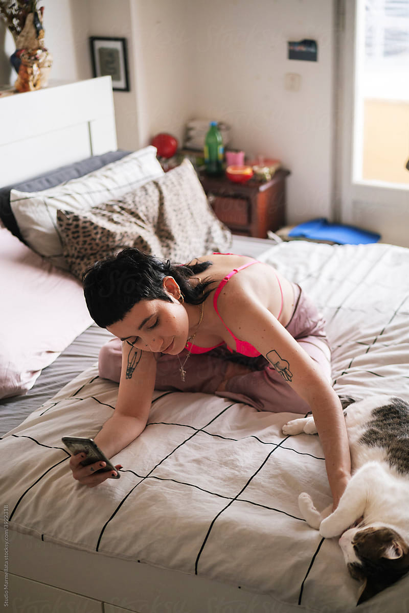 Glad alternative woman with smartphone stroking cute cat on bed