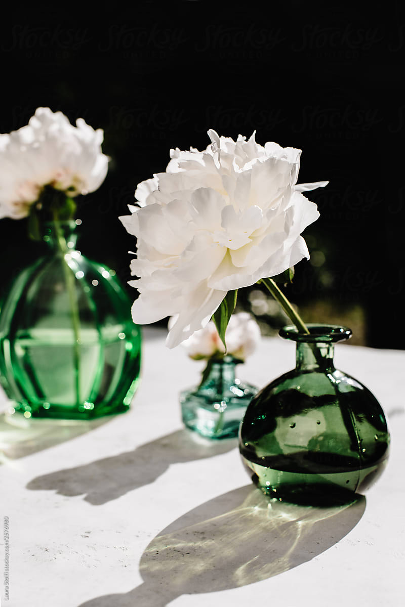 White peonies in green crystal bottles in direct sunlight