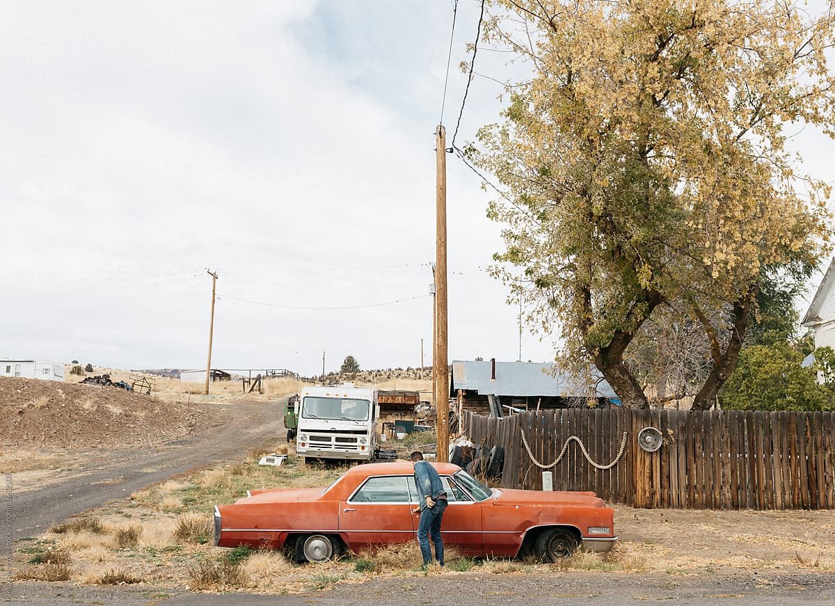 Young adult male in denim sits in front of broken down vintage car in rural town