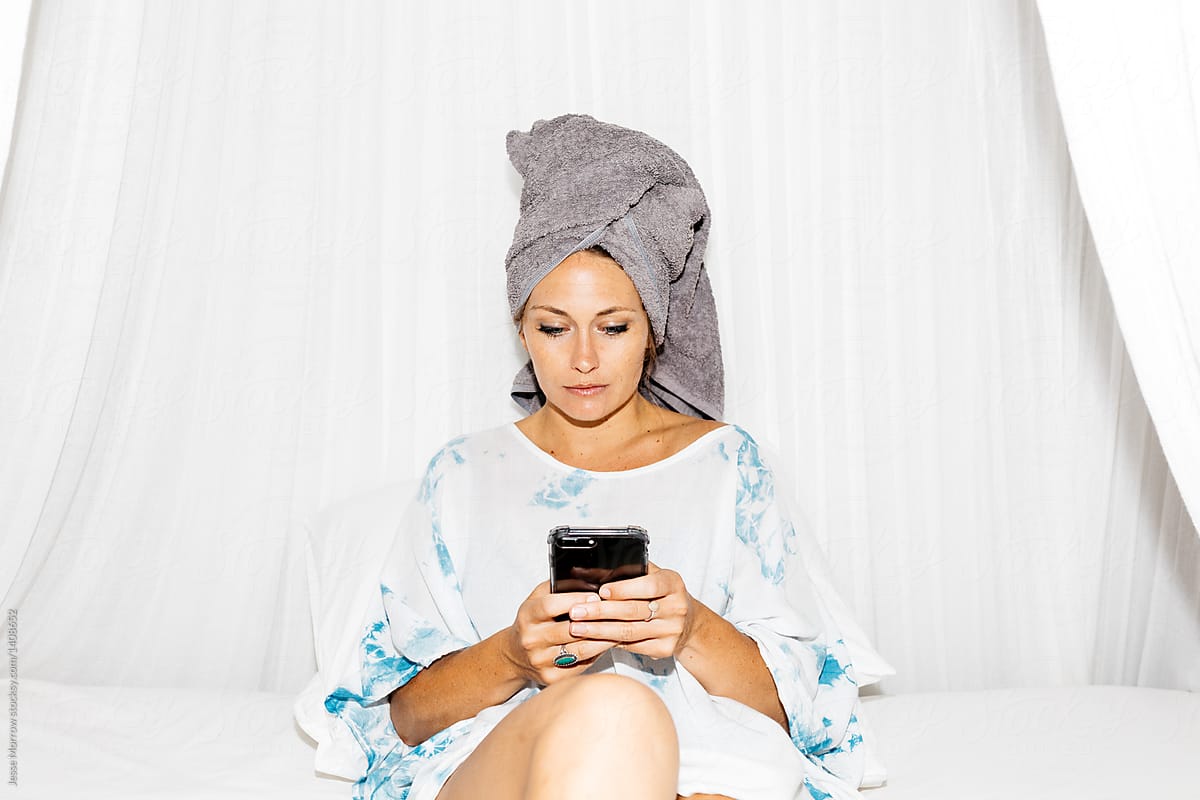 young adult woman sitting on bed with wet hair and towel on head looking at cell phone technology