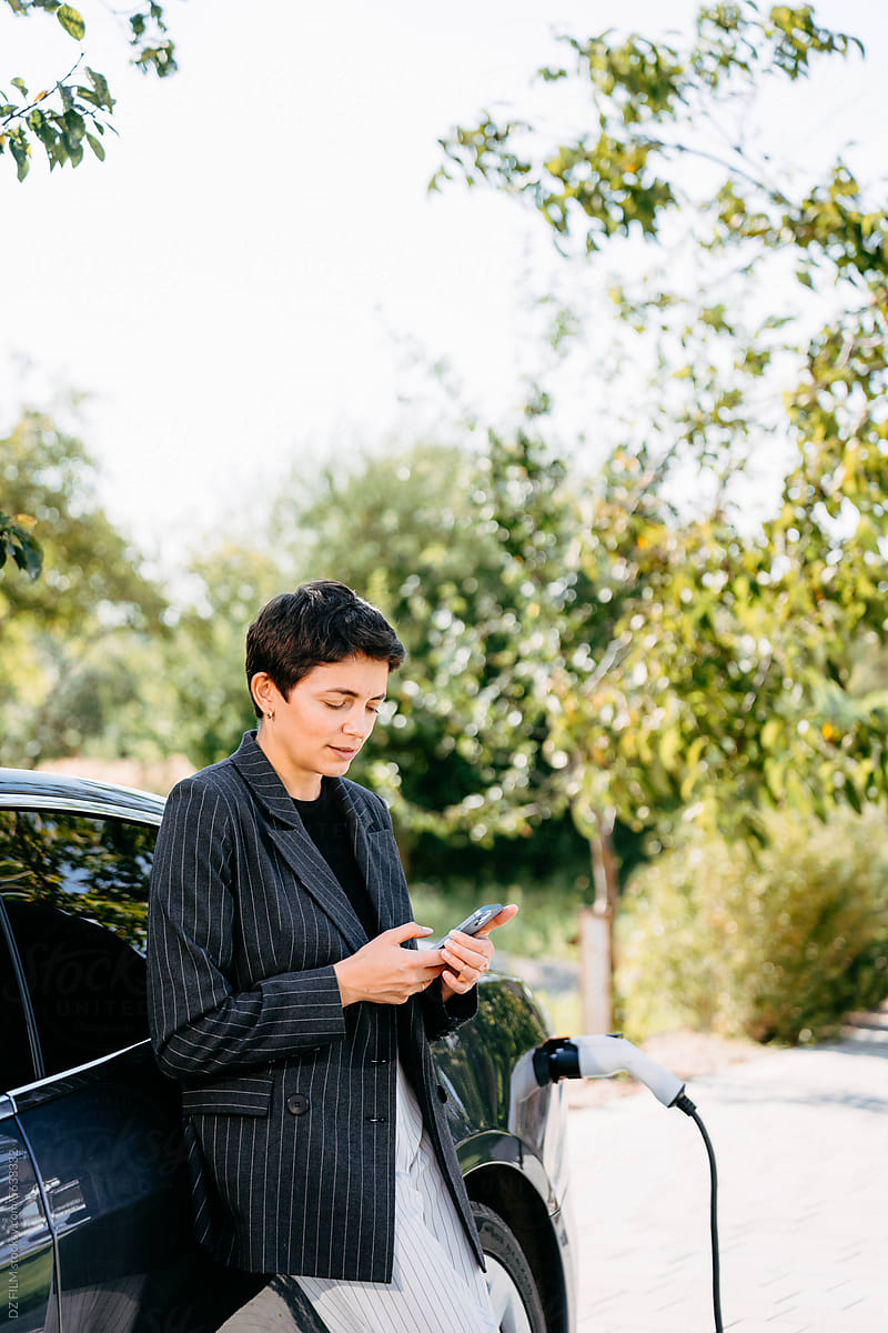 A woman uses a mobile phone standing near an electric car