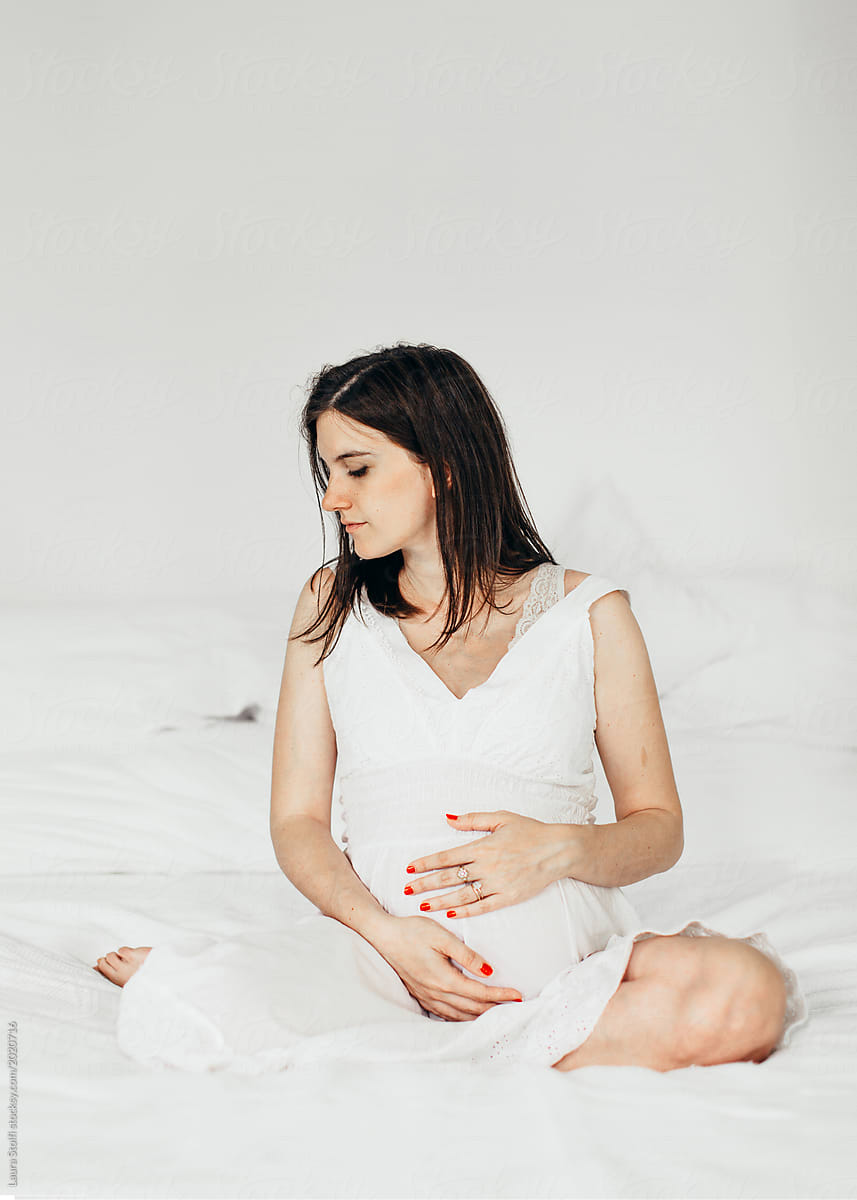 Beautfil woman in white sitting in bed caressing her pregnant belly