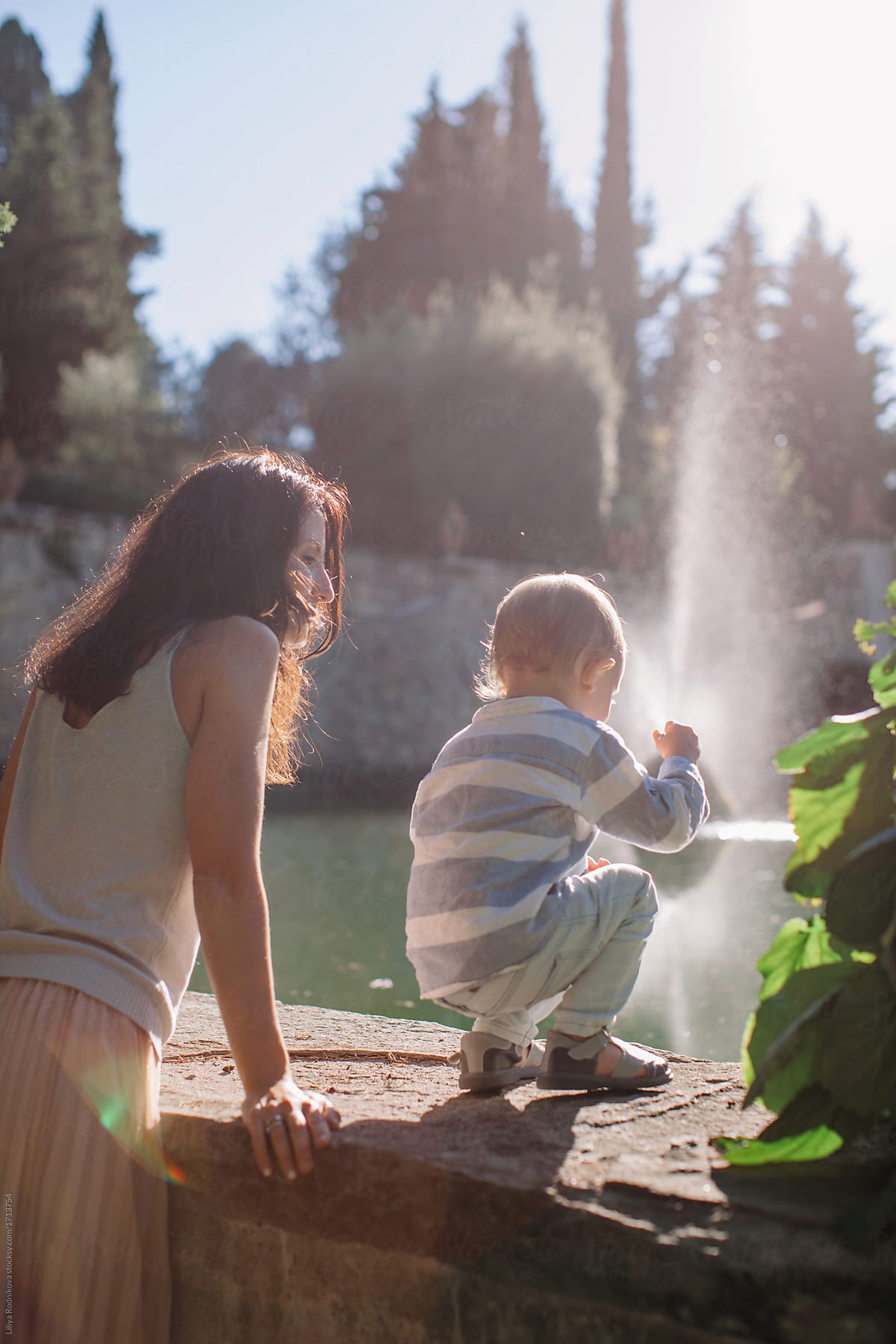 Back view portrait of mother and little son by the fountain in sunlight