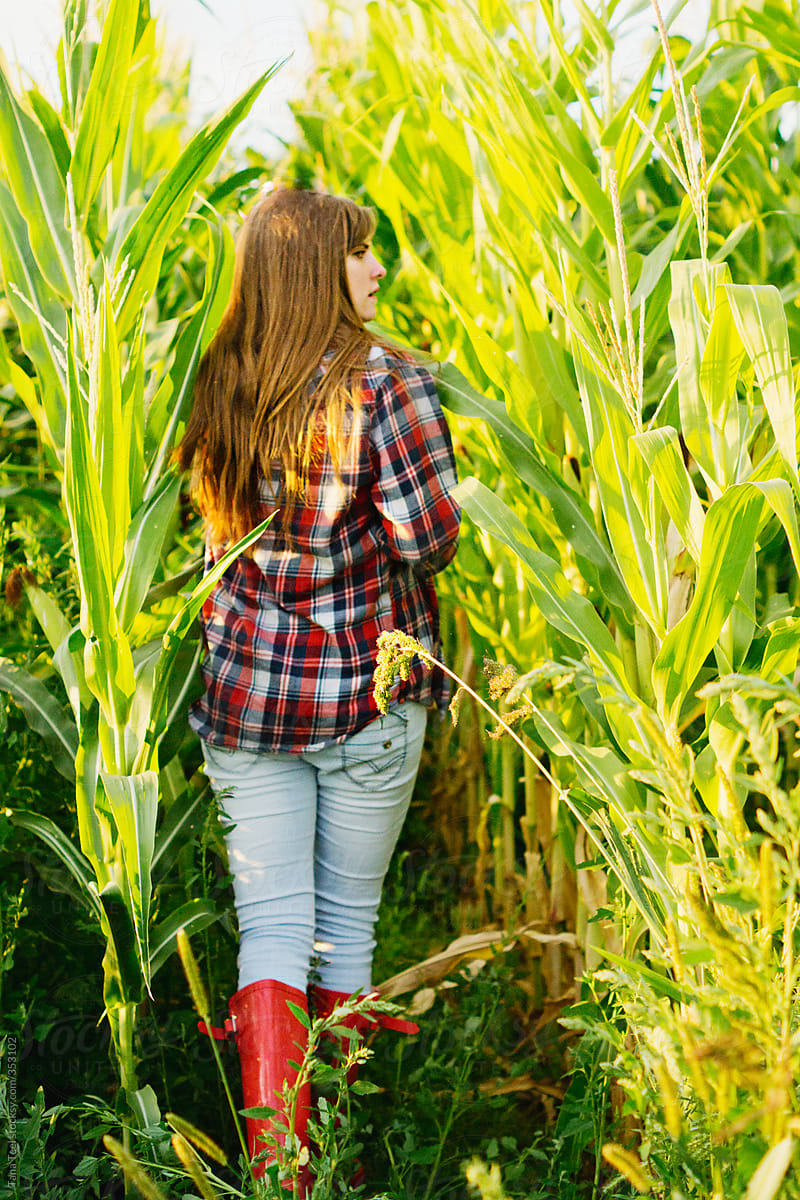 Young woman in flannel shirt and rubber boots walks into corn field