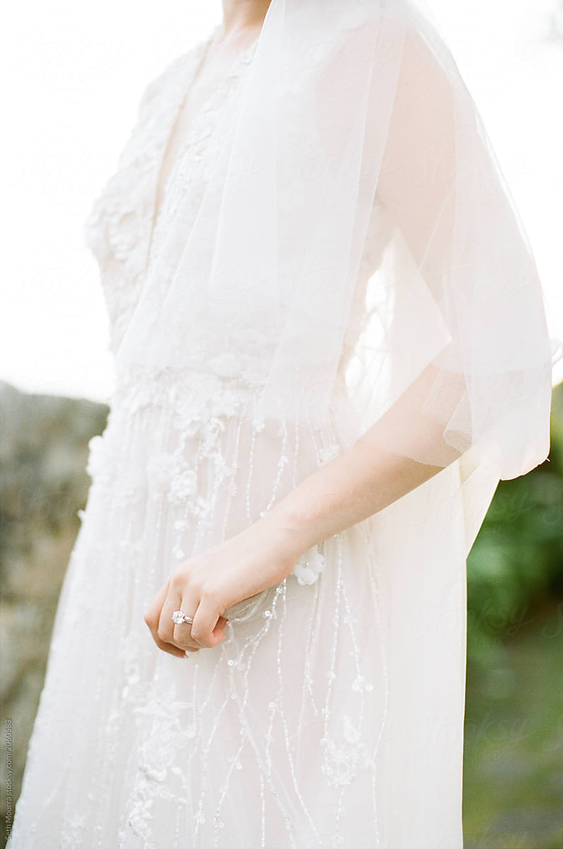 A bride in a delicate embroidered white wedding gown & veil draping gently holds her gown