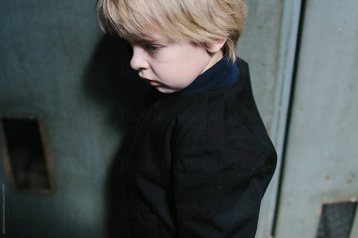Close-up profile of a blonde boy in a black jacket in a factory setting.