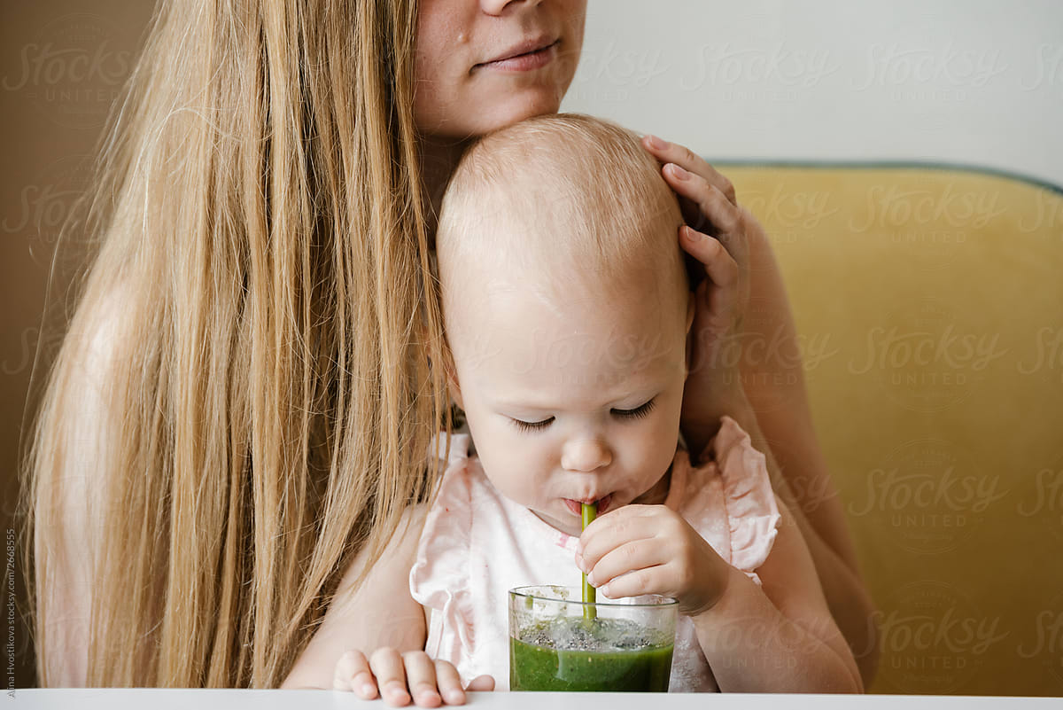 Baby girl with blue eyes drinking green smoothie with mom.