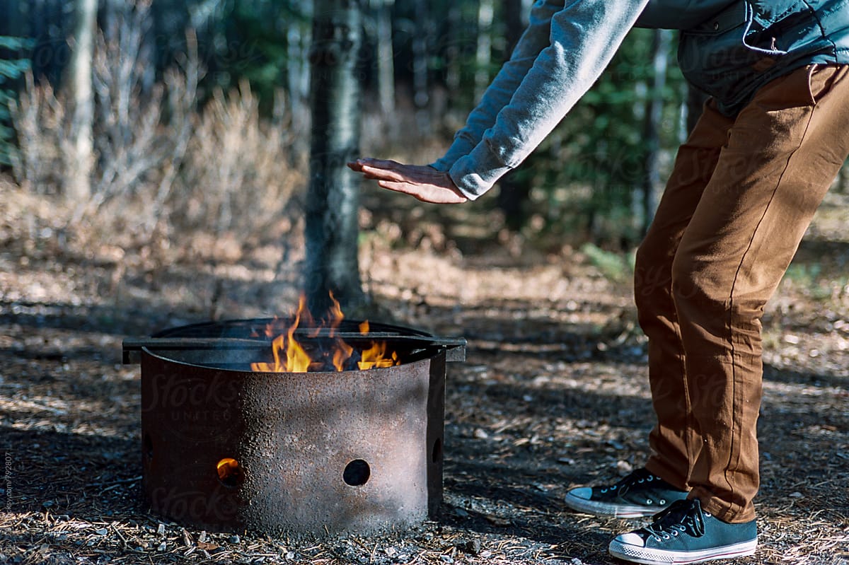 A man warms his hands over a fire in a camping fire pit.