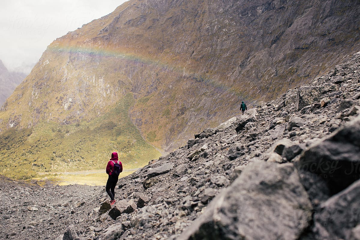A couple of hikers climbs a rocky mountain with a rainbow in the background.