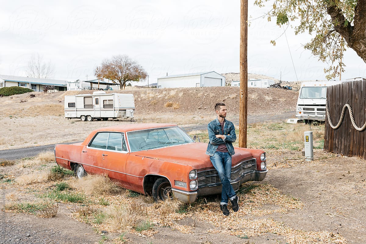 Young adult male in denim sits in front of broken down vintage car in rural town