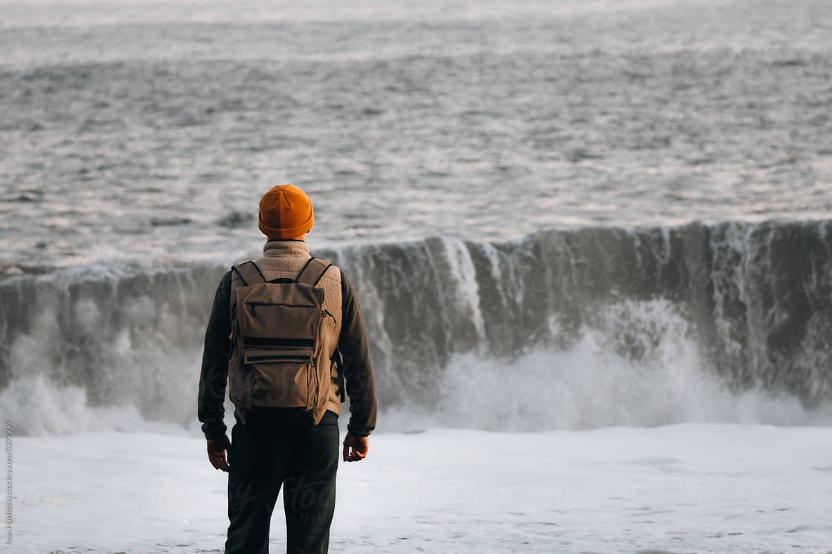 Man with backpack standing facing the big ocean waves from behind.