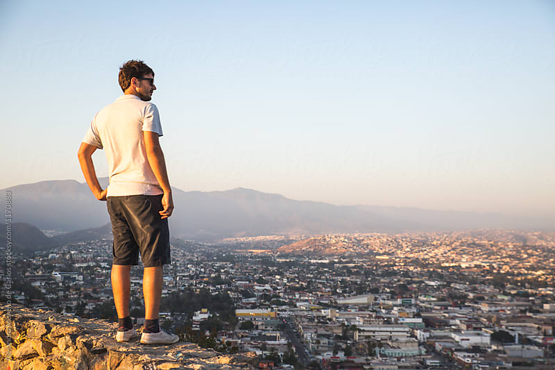 Young man standing on top of a wall overlooking a city at sunset from a viewpoint
