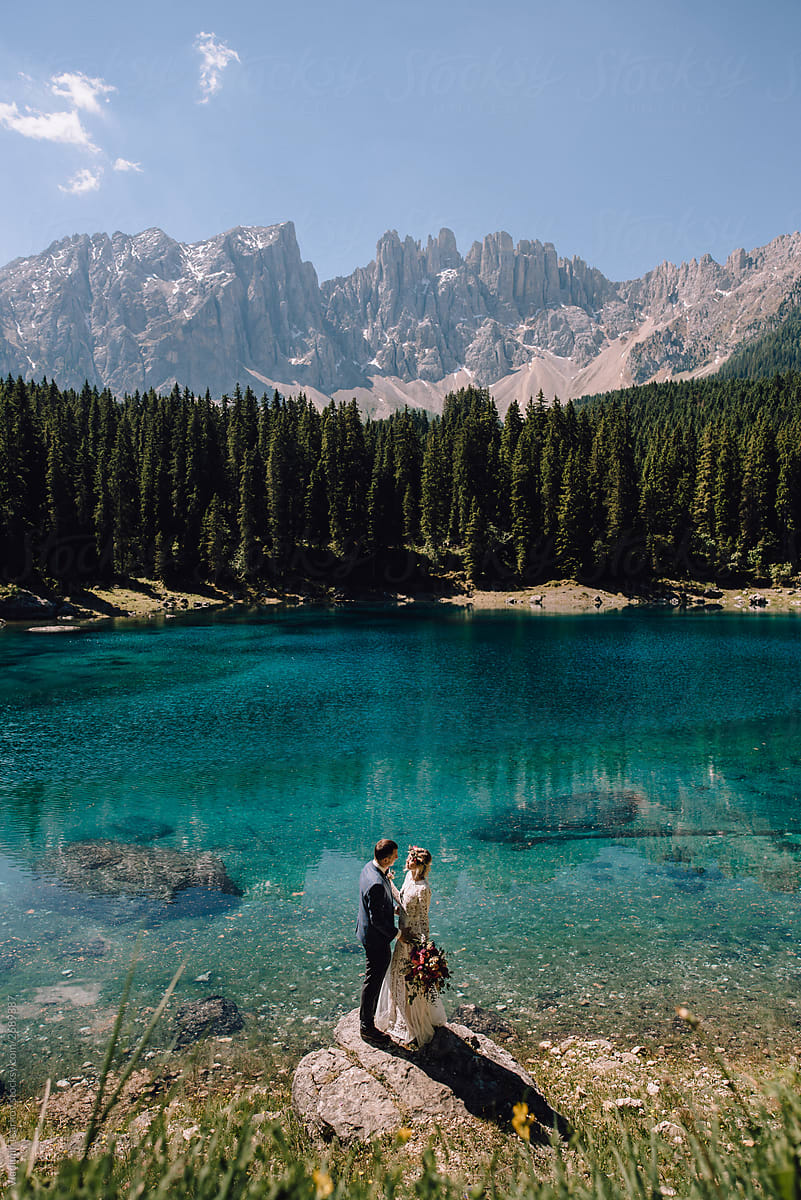 Beautiful couple in bridal wear standing on stone on mountain azure lake background