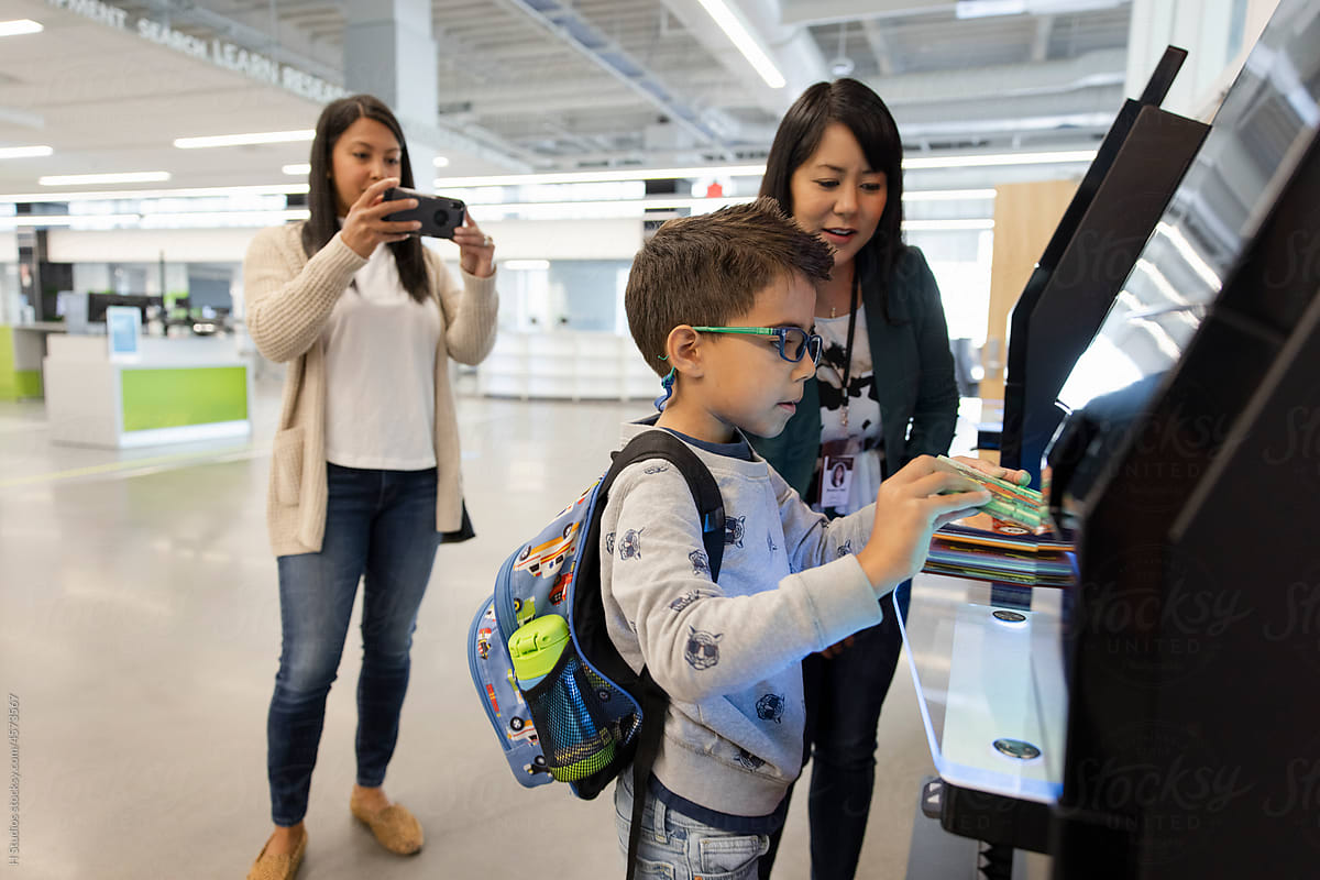 Mother photographing son using self checkout kiosk in library.