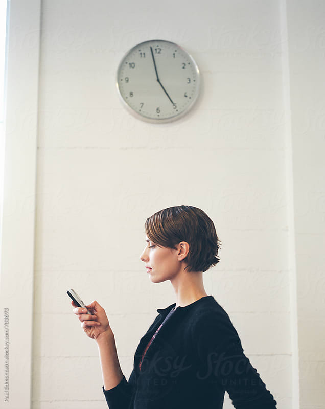 Female office worker looking at cell phone, clock above about to strike 5:00