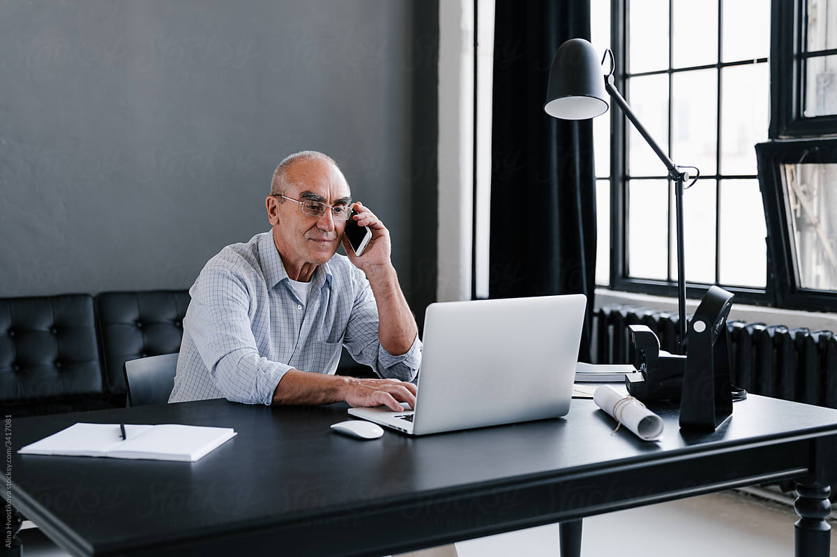 Busy Mature man looks at laptop at workplace while talking on phone
