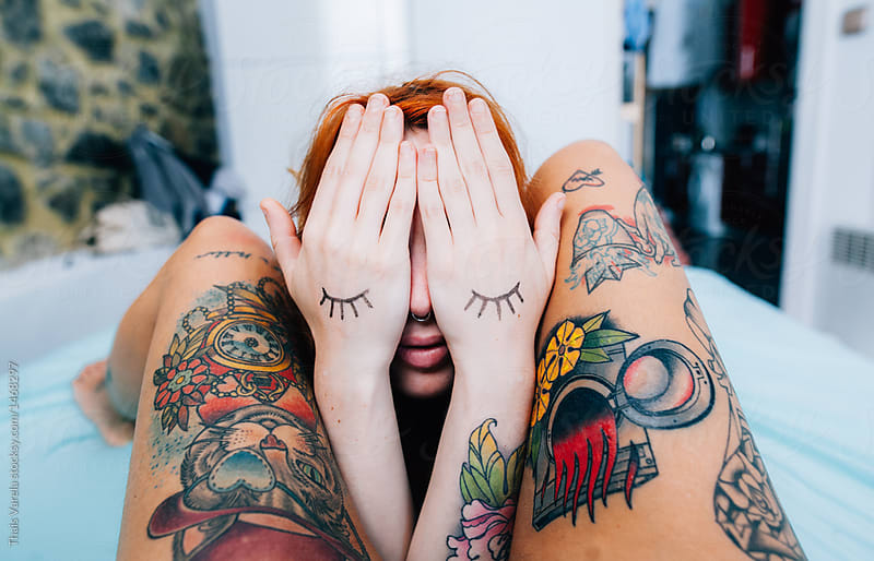 woman covering her face with drawn eyelashes on her hands between another womans tattoed legs