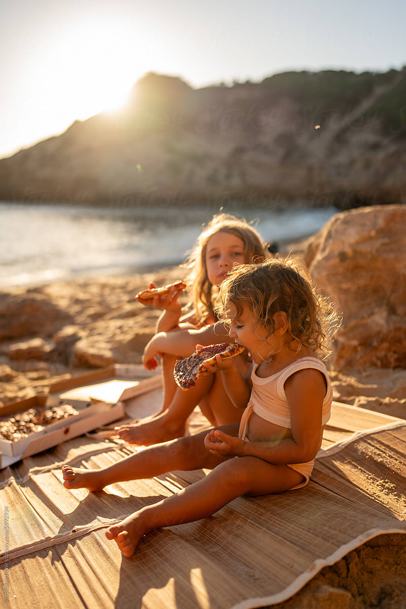 Siblings enjoying a pizza slice on the beach at sunset