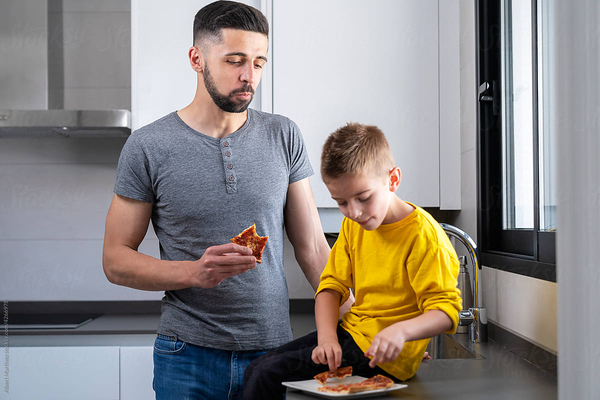 boy sitting on the worktop next to his dad eating pizza