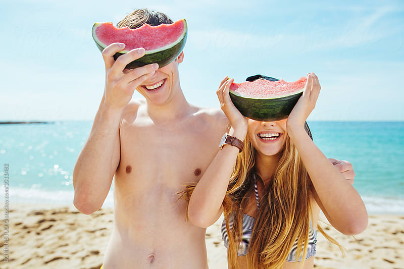 Smiling teenage couple hiding their eyes with a watermelon on the beach.
