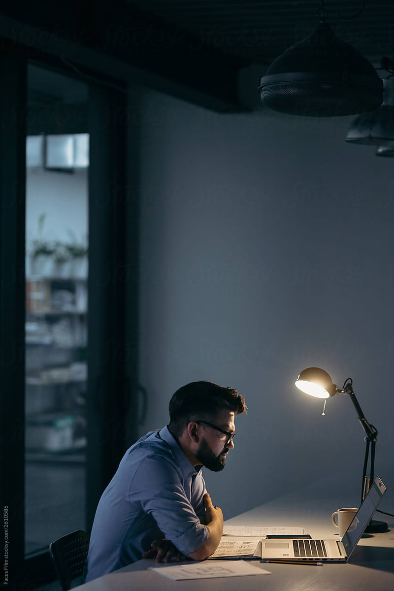 Caucasian man with beard and glasses sitting in dark office in front of laptop and table lamp. Night office work